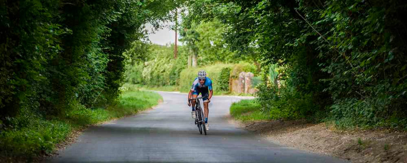 A cyclist in a blue and black outfit rides on a narrow, tree-lined country road. The greenery forms a natural archway overhead, creating a secluded and serene atmosphere. The road appears smooth and inviting for a peaceful ride.