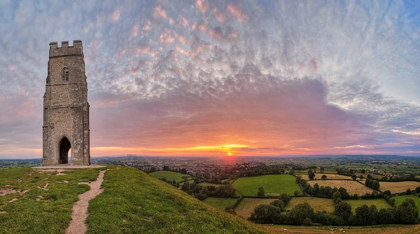 A historic stone tower stands atop a grassy hill during a picturesque sunset. The sky is painted with a blend of orange, pink, and purple hues, and the sun dips below the horizon. Rolling green fields and scattered trees stretch out in the distance.