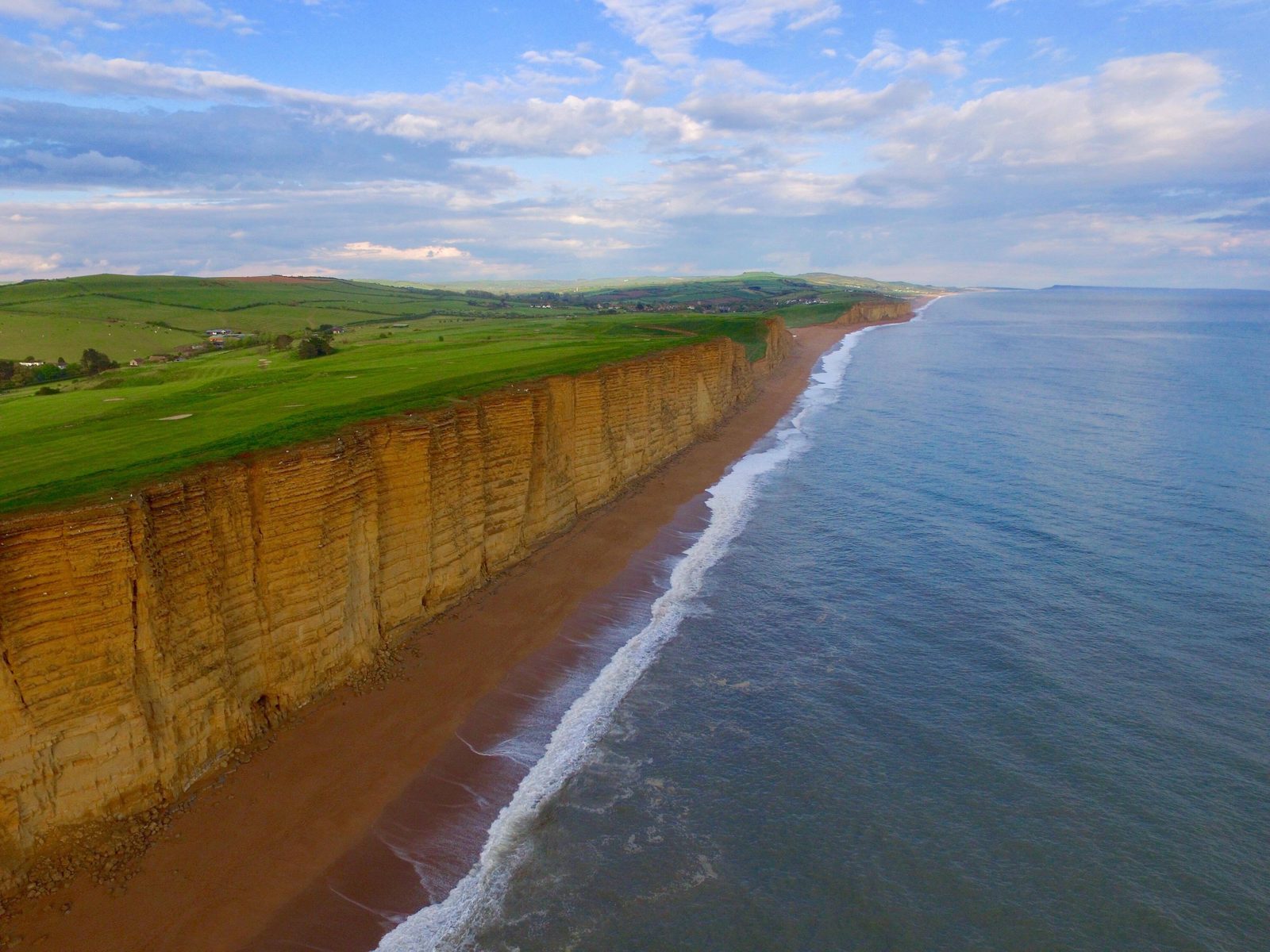 Aerial view of sandstone cliffs along a beach with waves gently lapping the shore. Lush green meadows blanket the top of the cliffs, extending into the distance. The sky is partly cloudy with patches of blue, enhancing the coastal landscape’s beauty.