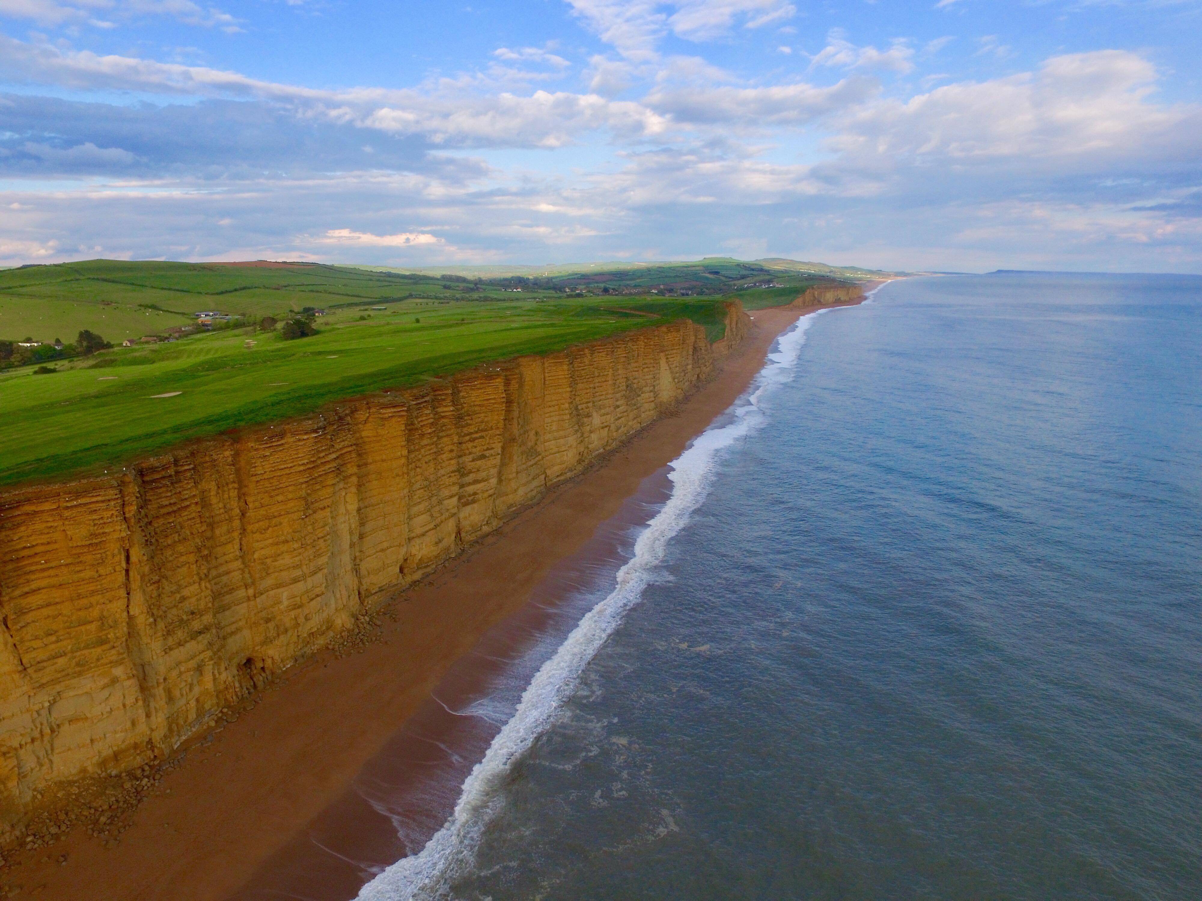 Aerial view of sandstone cliffs along a beach with waves gently lapping the shore. Lush green meadows blanket the top of the cliffs, extending into the distance. The sky is partly cloudy with patches of blue, enhancing the coastal landscape’s beauty.