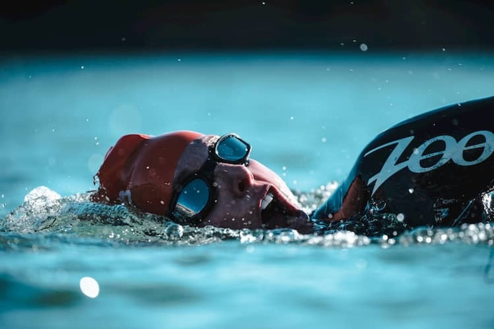 Close-up of a swimmer in a wetsuit and red swim cap, swimming freestyle in open water. The swimmer is wearing goggles and turning their head to the side to breathe. The water surface is calm with slight ripples, and drops of water are visible around the swimmer's face.