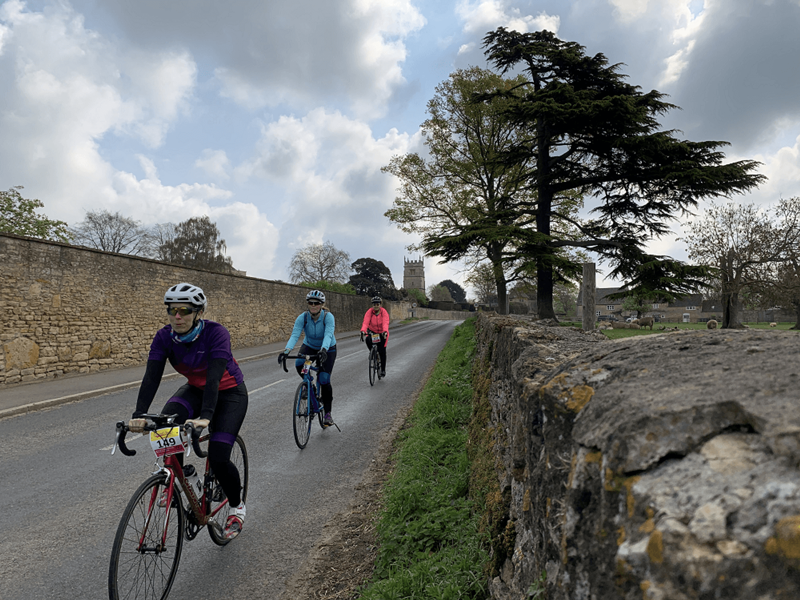 Three cyclists ride on a paved, rural road in light gear, with numbers on their bikes. They pedal past a stone wall and tall trees, with a church tower visible in the background. The sky is partly cloudy.
