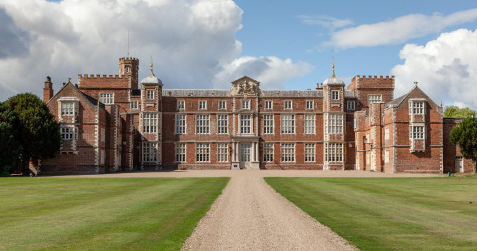 A grand, historic mansion with red brick walls and numerous windows, featuring a central tower, chimneys, and ornate architecture. It is set in a spacious green lawn under a partly cloudy sky. A gravel pathway leads directly to the entrance.
