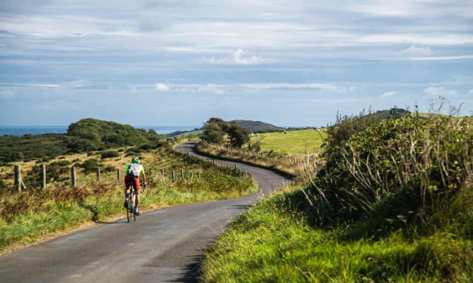 A cyclist in a red and green outfit rides on a narrow, winding road through a lush, green countryside with hills and trees under a partly cloudy sky.