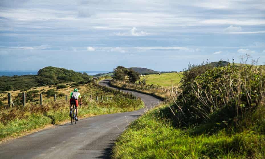 A cyclist in a red and green outfit rides on a narrow, winding road through a lush, green countryside with hills and trees under a partly cloudy sky.