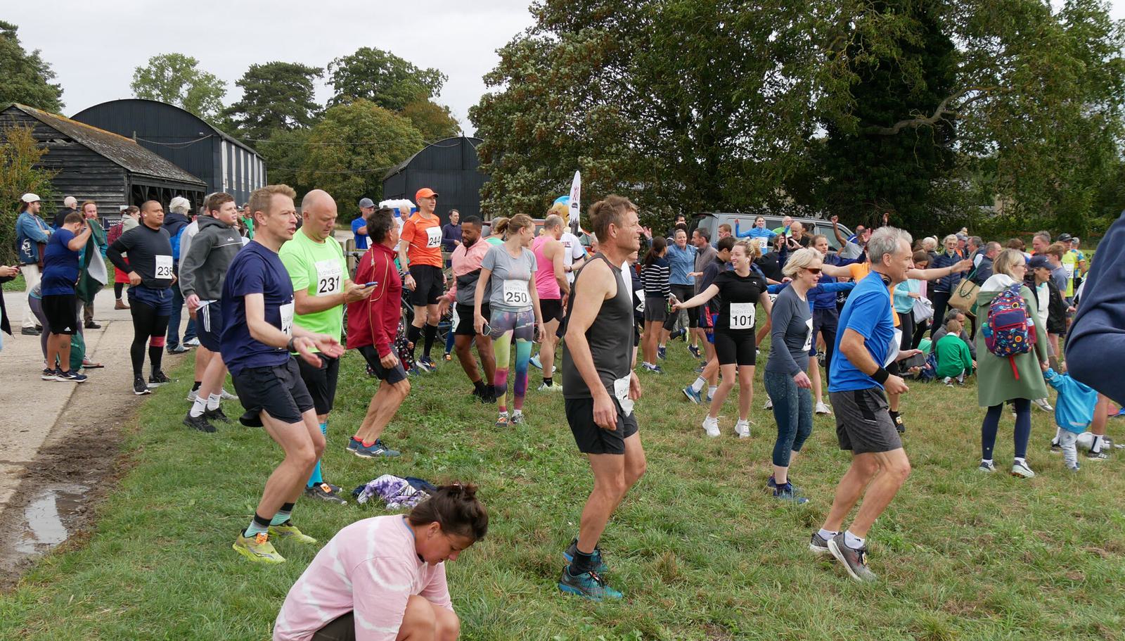 A diverse group of people are gathered outdoors for a race event. Some participants are jogging in place and stretching, wearing running gear with visible race bibs. Spectators, including families and pets, mingle nearby on a grassy area under a cloudy sky. .