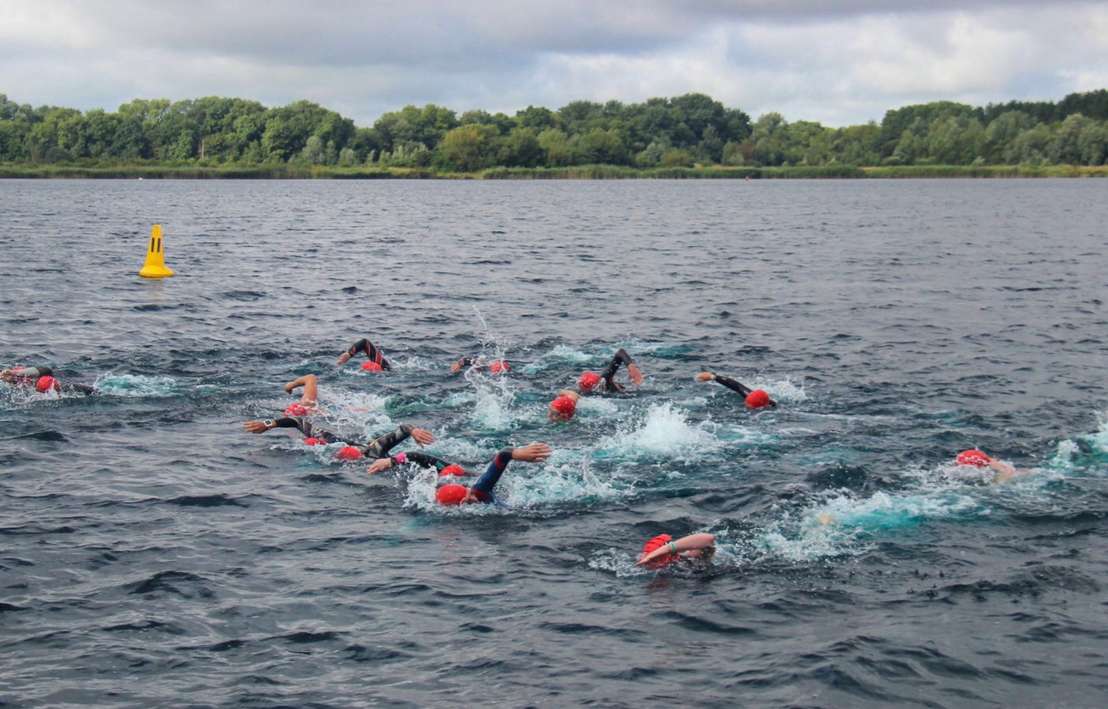 A group of swimmers wearing wetsuits and red swim caps are competing in open water under a cloudy sky. They are swimming close together in a lake surrounded by lush green trees. A yellow buoy is visible in the distance, marking part of the course.
