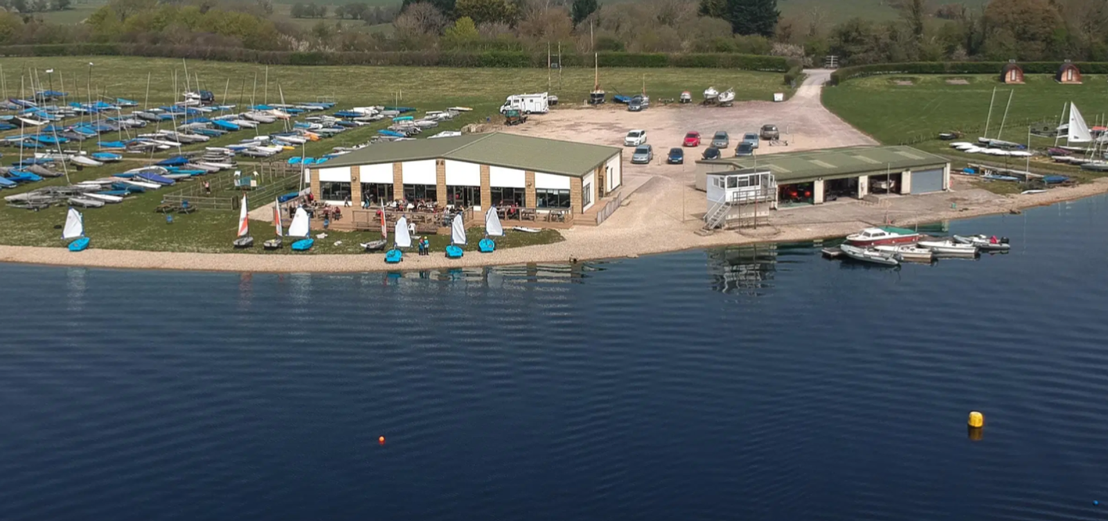 Aerial view of a lakeside sailing club with several boats on the shore and docked on the water. The club features a central building with a green roof and adjacent parking area. Numerous sailboats are arranged on the grass and in the water, ready for use.