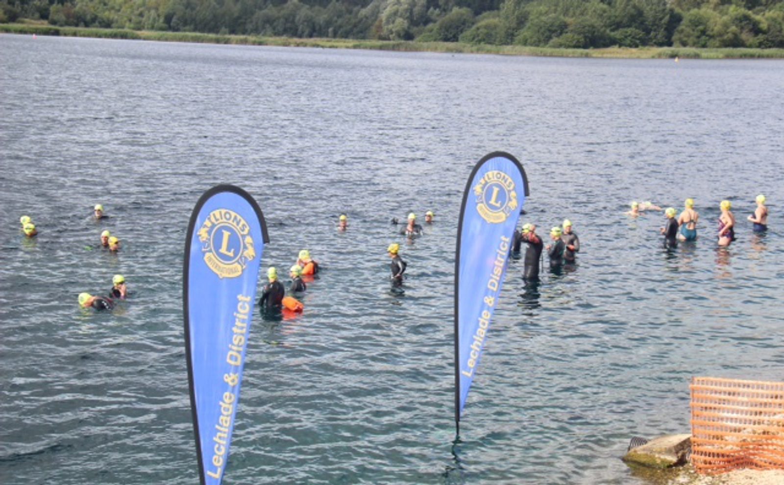 Swimmers wearing wetsuits and yellow swim caps participate in an open water event in a lake, with blue Lechlade & District Lions Club banners displayed near the shore. The background features greenery and trees.