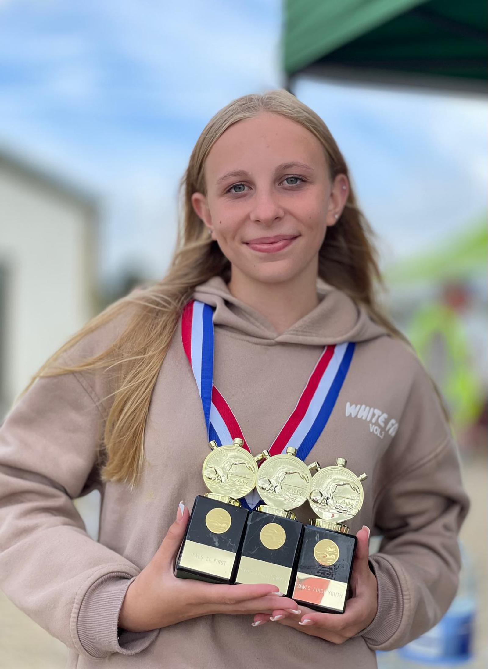 A young girl smiles while holding three trophies with medals around her neck. She is wearing a beige hoodie and standing outdoors under a green canopy. The background is blurred, highlighting her achievements.
