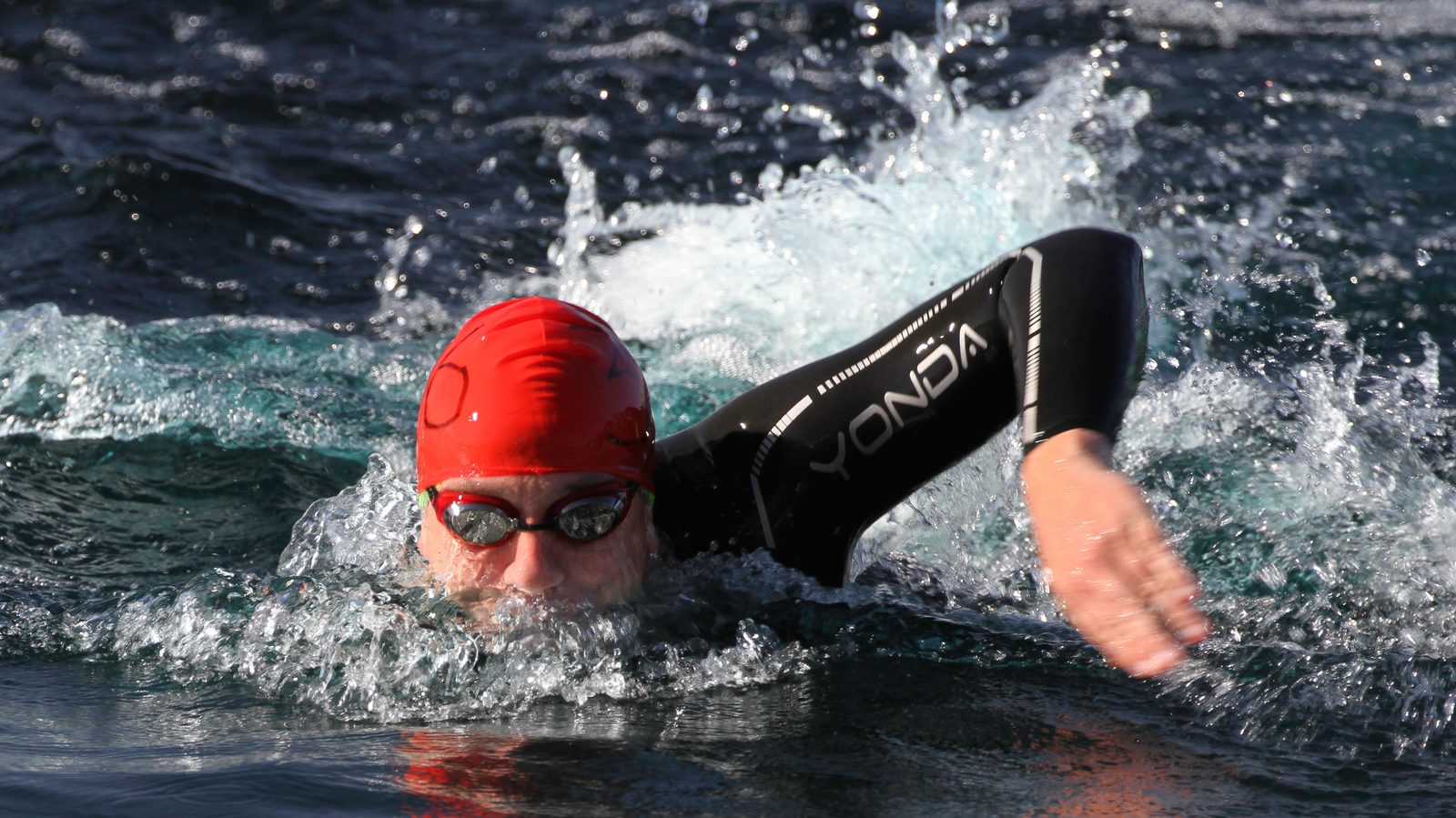 A person wearing a red swim cap, goggles, and a black wetsuit swimming freestyle in open water. Waves ripple around them as they move forward, with their arm extended in a stroke.