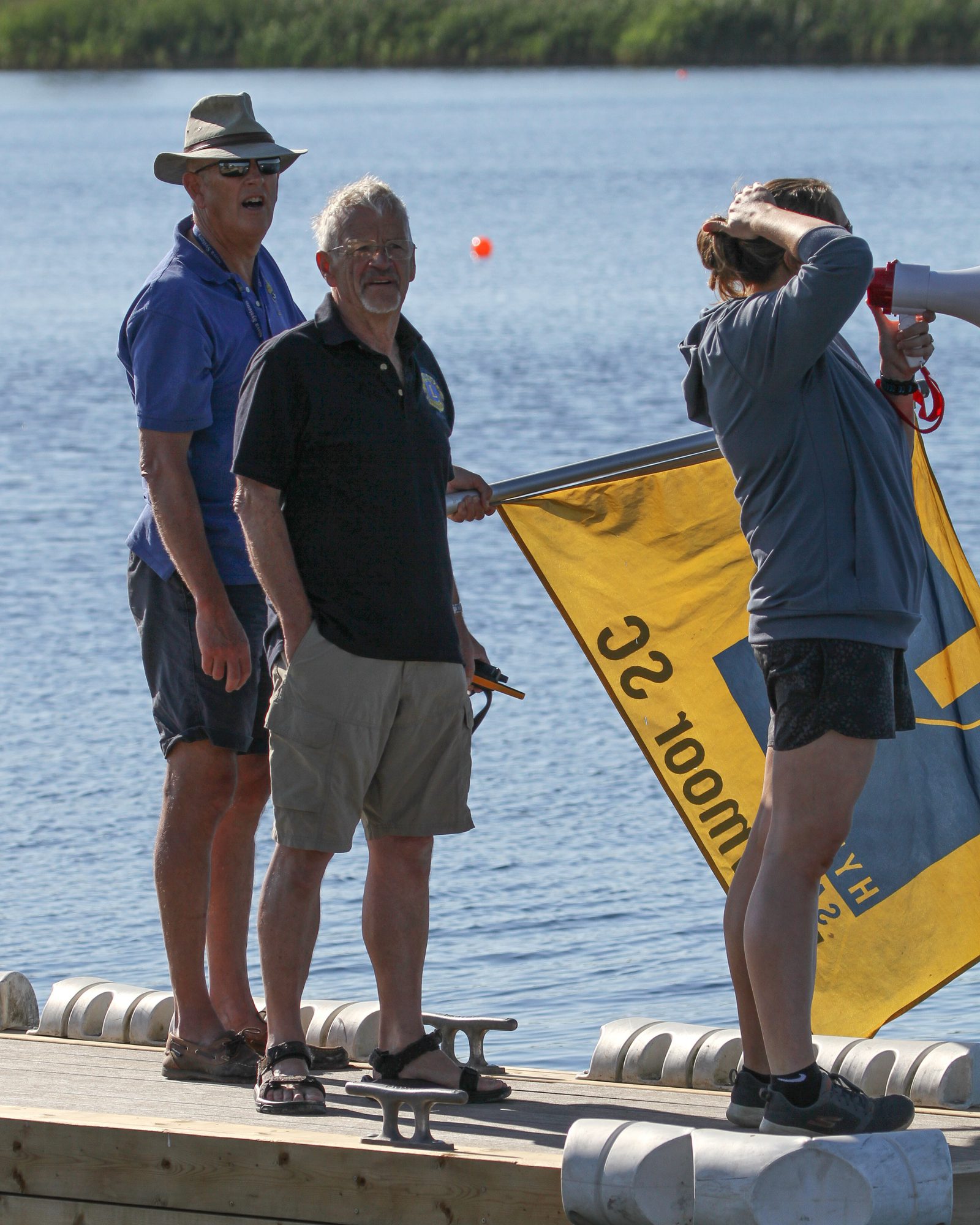 Three people stand on a dock by the water. Two men are wearing hats and casual clothing, one in blue and one in black. The third person, wearing a gray shirt and holding a megaphone, is raising a yellow and blue flag. The background shows calm water and green foliage.