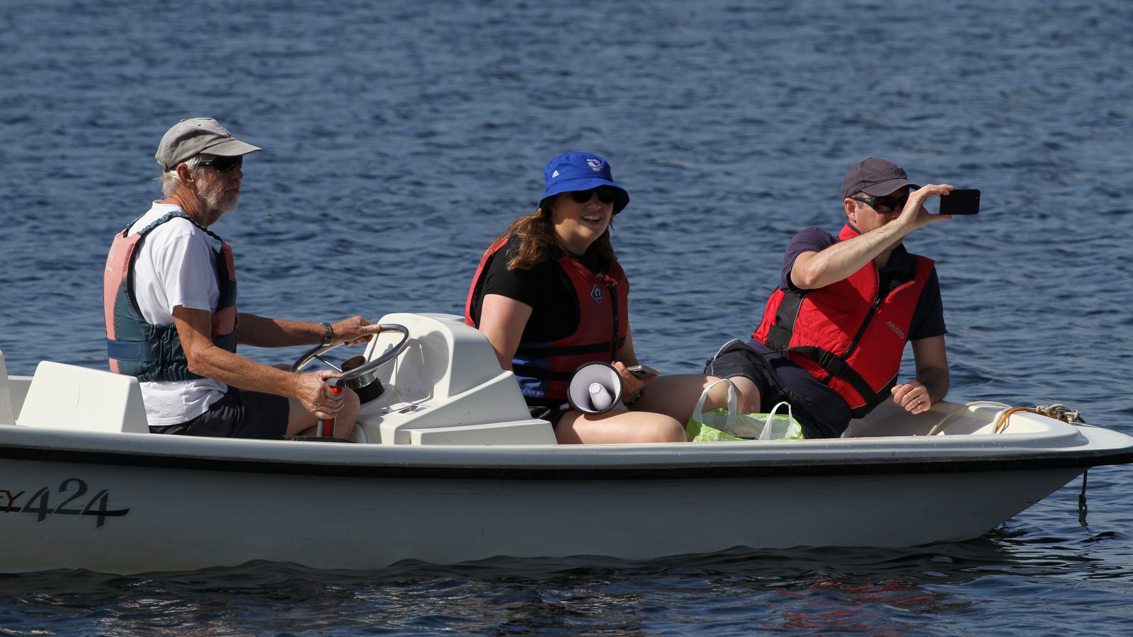 Three people are seated in a small white boat on a body of water. The person on the left is steering, the one in the middle is smiling and holding a cup, and the person on the right is holding up a phone, possibly taking a photo or video. All are wearing life jackets.