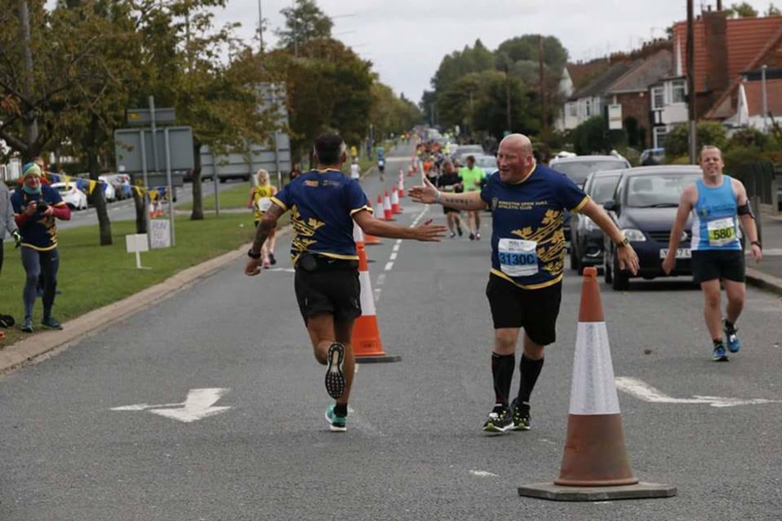 Two runners participate in a road race, high-fiving each other as they pass. They are wearing race bibs and blue shirts with yellow designs. Other runners are visible in the background. The road is lined with traffic cones and spectators watch from the sides.