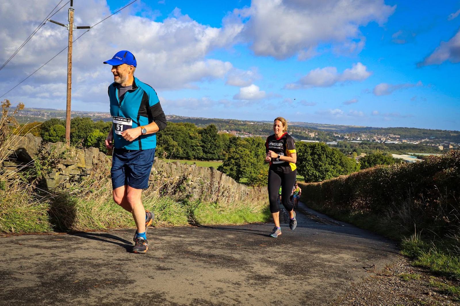 Two runners participating in a race are on a scenic outdoor path, with a man in a blue shirt and blue cap running ahead and a woman in a black shirt following. The background features a vibrant green landscape and a blue sky with scattered clouds.