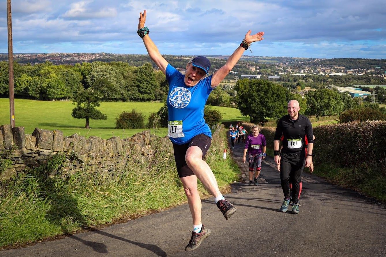 A person in a blue shirt and black shorts is joyfully jumping in the air during a race on a scenic rural road with lush green fields and trees in the background. Other participants, including a person in a black outfit, walk behind them under a partly cloudy sky.