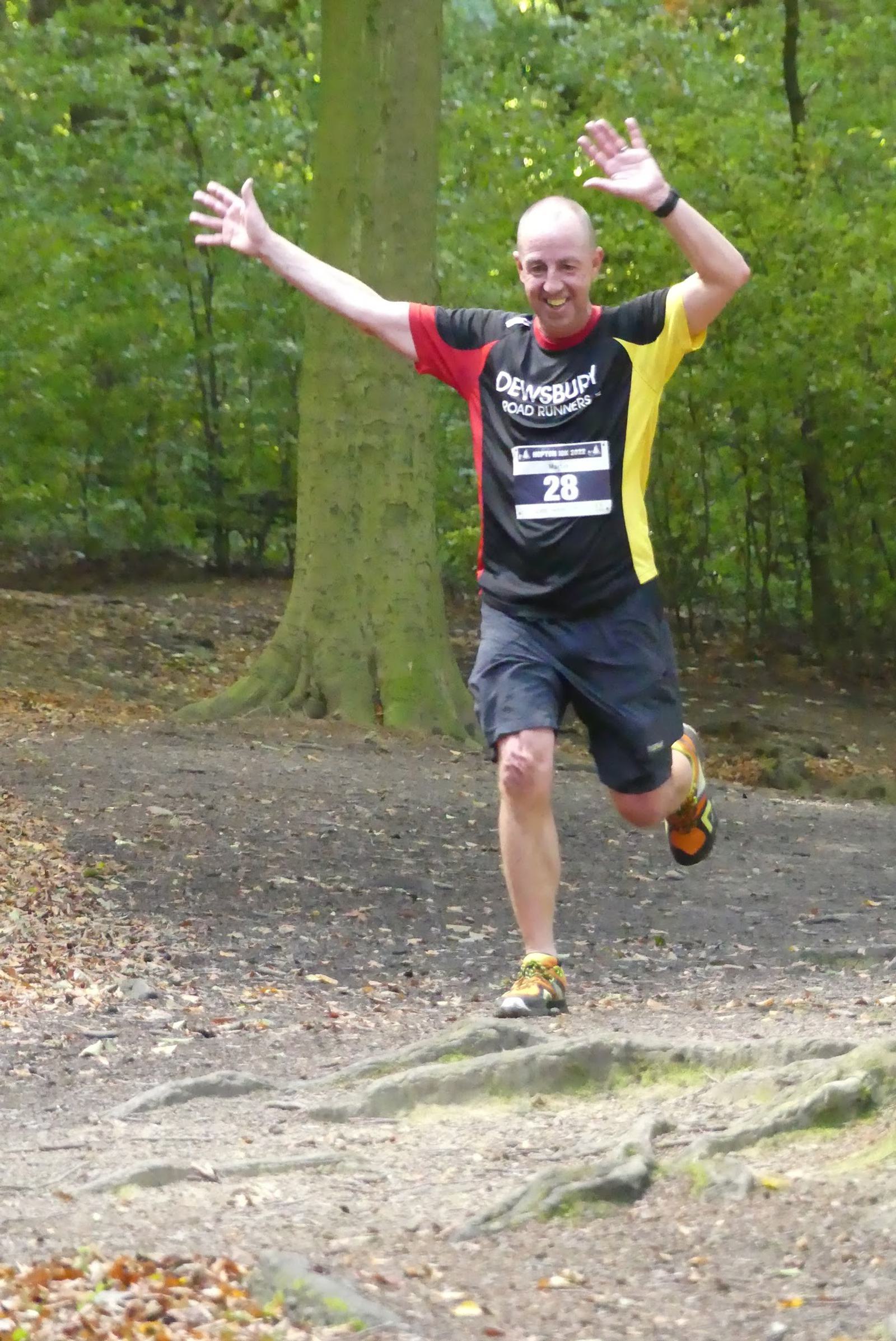 A man is running on a forest trail with his arms raised in celebration or joy. He is wearing a black and red running shirt, black shorts, and running shoes. He has a race number 28 pinned to his shirt. The background is full of green trees and a dirt path.