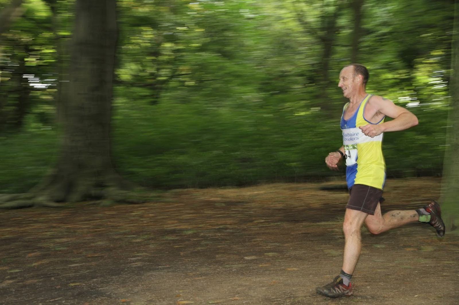 A man in a blue and yellow tank top and brown shorts is running on a dirt path in a forest. The background is a blur of green trees, emphasizing his speed. He appears focused and is mid-stride, with one foot off the ground.