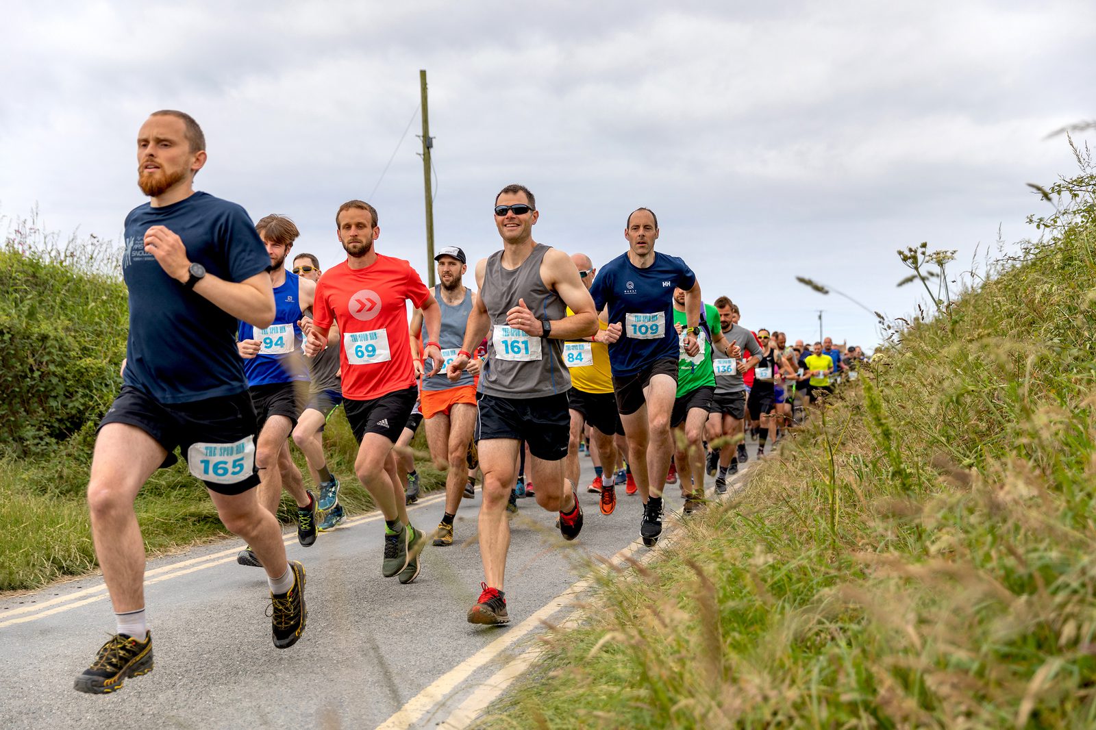 A group of runners participates in a race on a country road. The runners wear various colored athletic gear and numbered bibs. Lush green vegetation flanks the road, and the sky is overcast. The lead runner wears a blue shirt and black shorts, number 165.