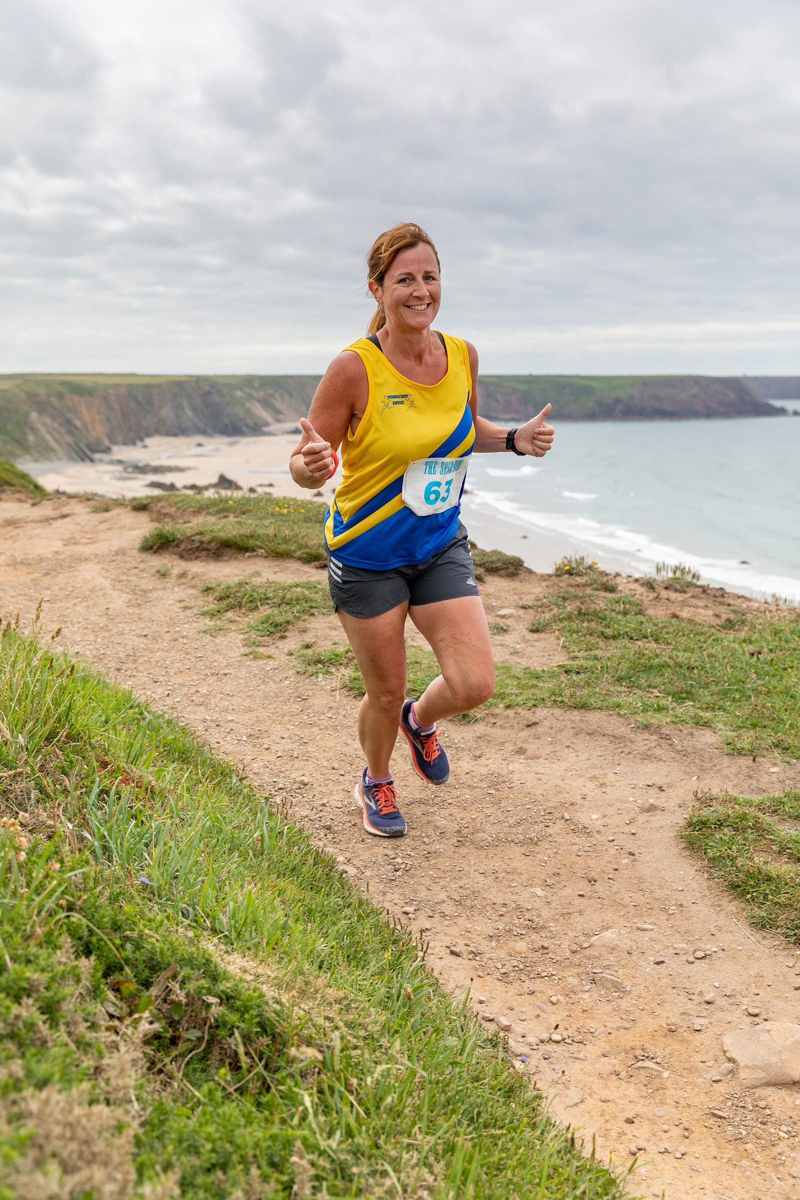 A woman in athletic gear is running on a coastal trail, with the ocean and cliffs in the background. She is smiling and giving two thumbs up, wearing a yellow and blue tank top, grey shorts, and blue running shoes. It's a cloudy day.