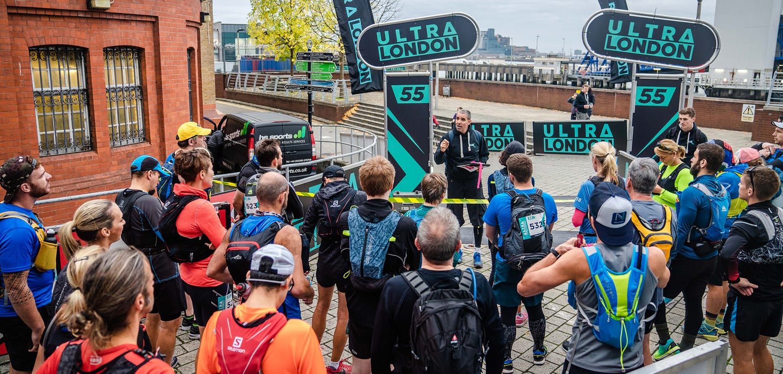A group of runners gathers around a man giving a pre-race briefing at the starting line of the Ultra London race. The runners are wearing colorful athletic gear and race bibs. Large banners with the race name "Ultra London" are visible in the background.
