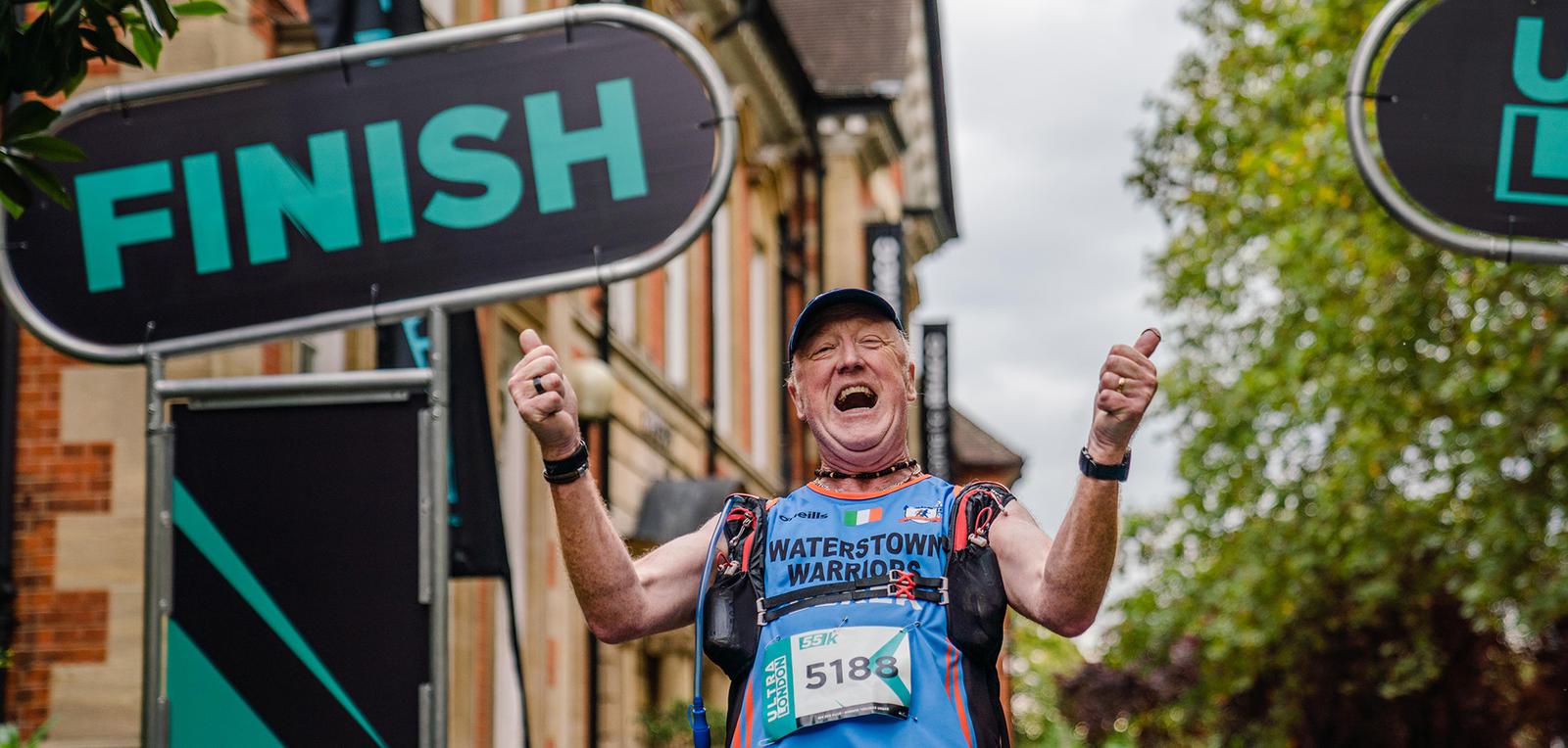 An enthusiastic runner crosses the finish line, raising both thumbs up in celebration. He is wearing a blue running outfit with "Waterstown Warriors" printed on the front and a race bib numbered 5188. Tall buildings and trees line the background, under cloudy skies.