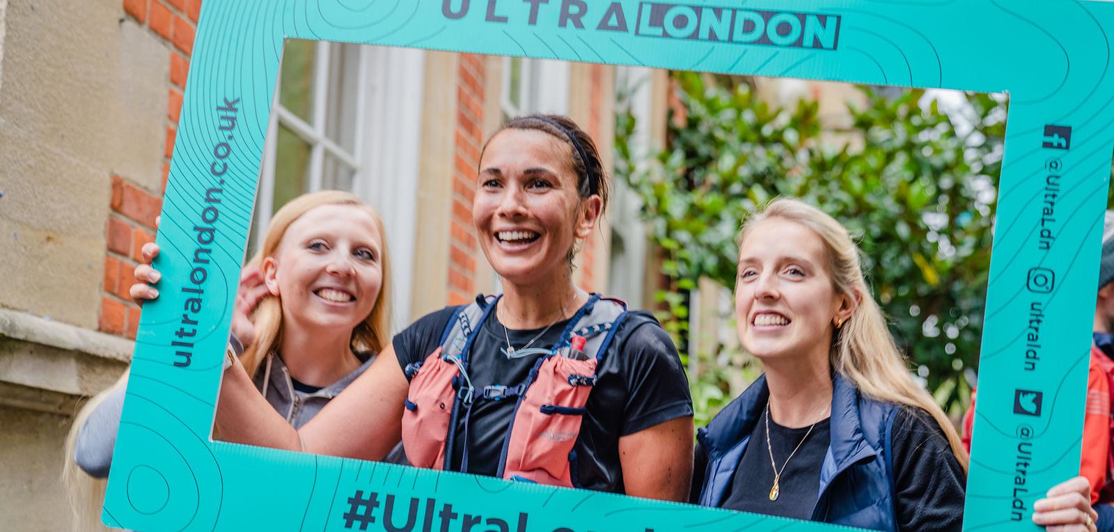 Three smiling women are framed by a large teal-colored sign with the text “ULTRA LONDON” around the top and social media handles on the side. They appear happy and posed for a photo, likely after participating in an event. The background features buildings and greenery.