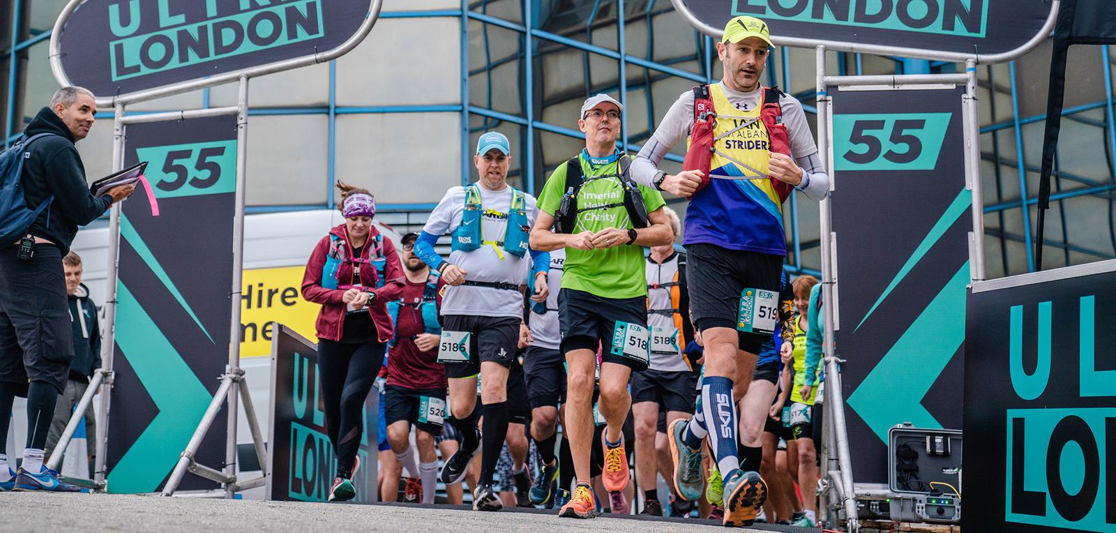 A group of runners, wearing sports gear and race numbers, start a race under a banner that says "Ultra London." Some runners wear hydration packs and headbands. The background shows a building's glass facade and additional race signage.