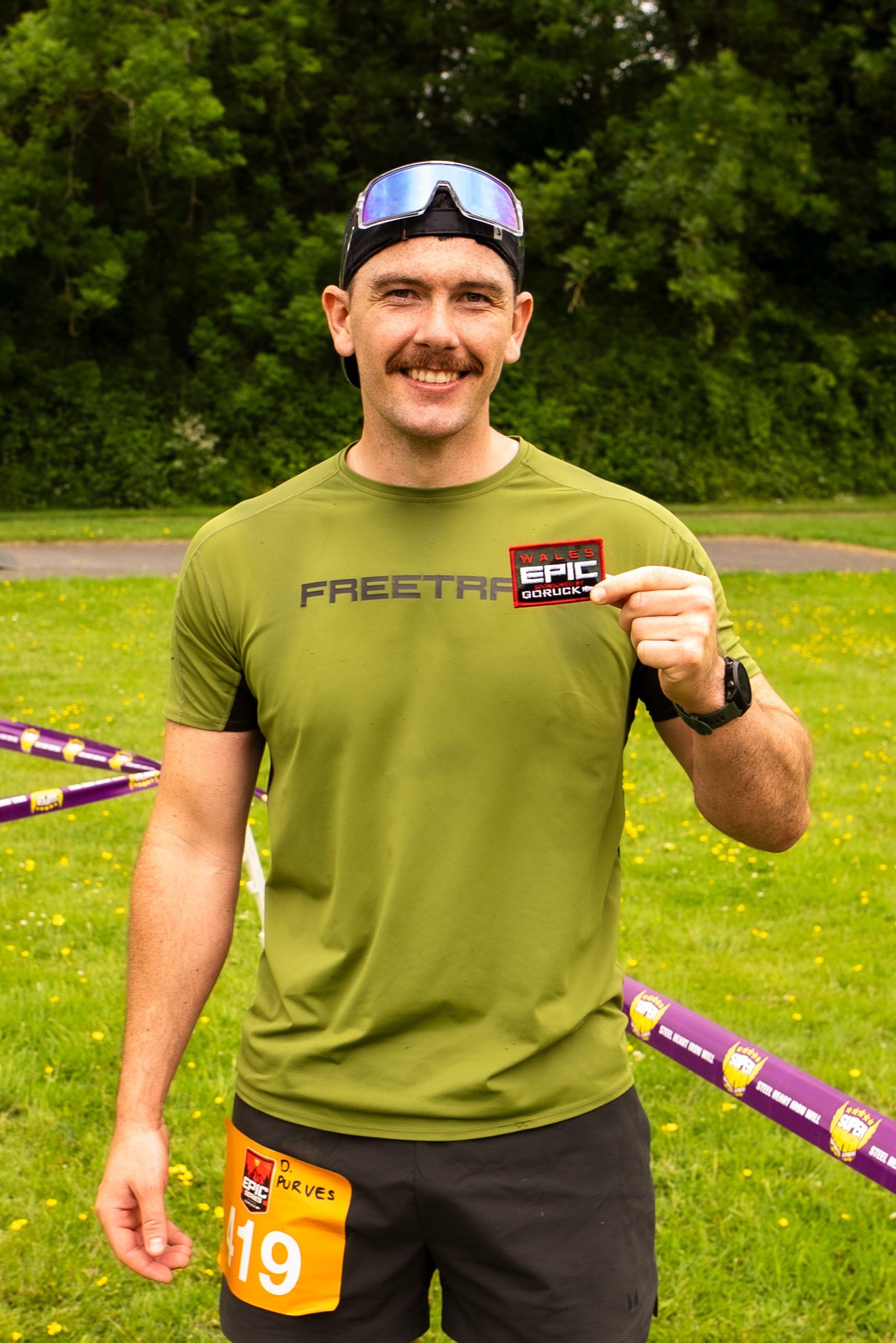 A smiling man with a mustache, wearing sunglasses on his head, a green T-shirt, and race bib 419, stands outdoors holding a "Go Ultra EPIC" badge. Green grass and trees are visible in the background.