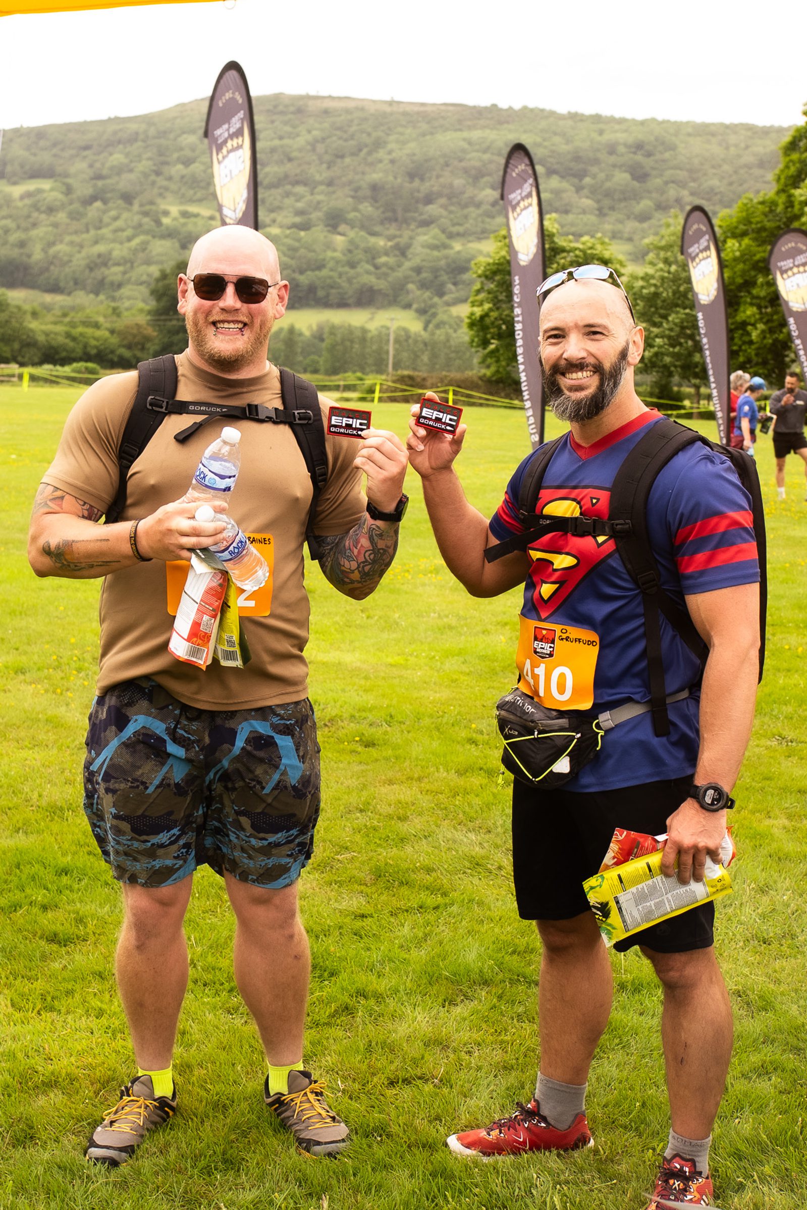 Two smiling men stand on grass at an outdoor event, holding up energy bars. One wears camouflage shorts and sunglasses; the other wears a Superman T-shirt and race bib. Flags and green hills are visible in the background.