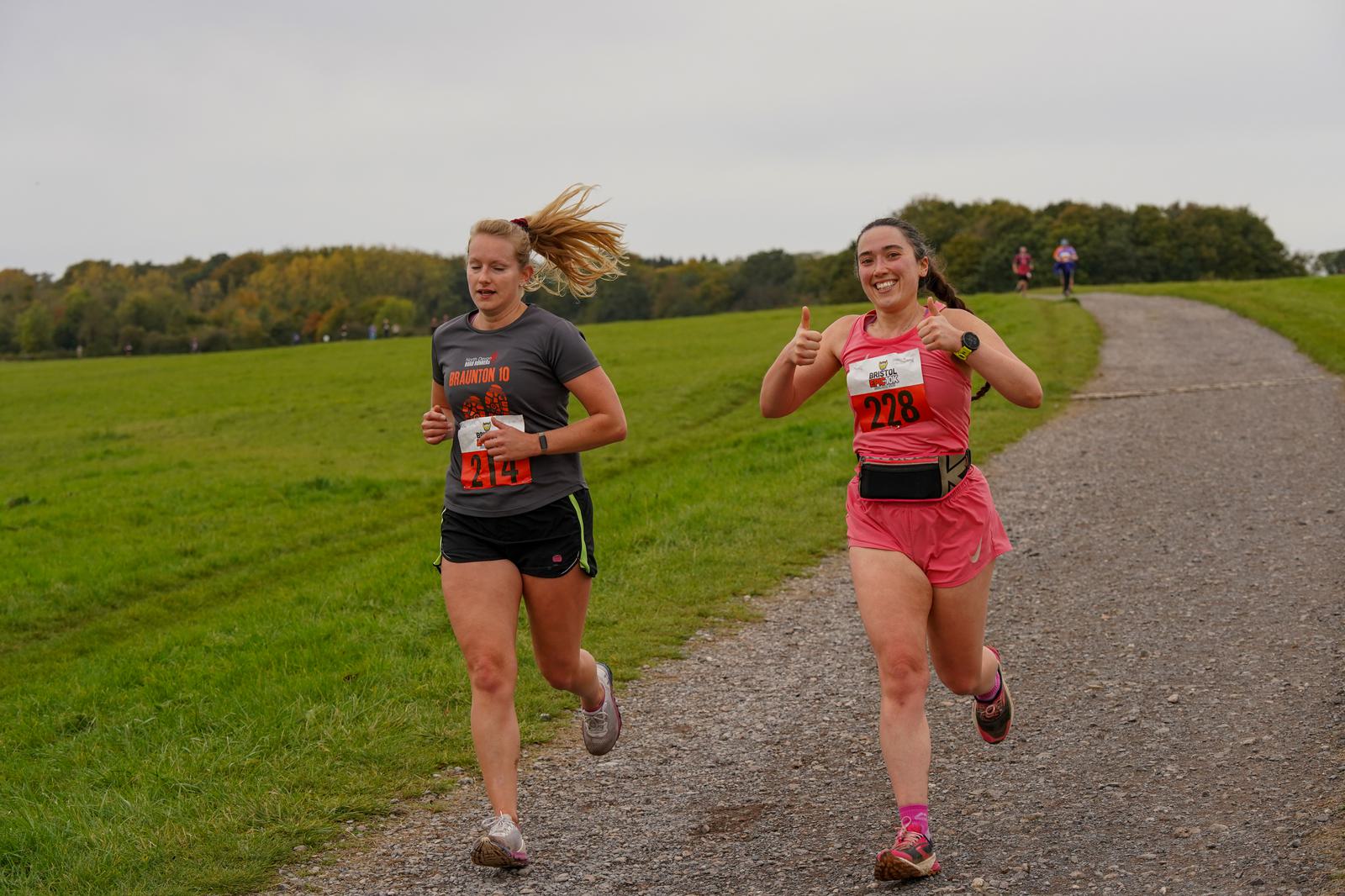 Two women are running on a gravel path through a green park. One wears a bib numbered 428 and has her hair in a ponytail. The other, wearing bib number 228, smiles and gives a thumbs-up. Trees are visible in the background under a cloudy sky.