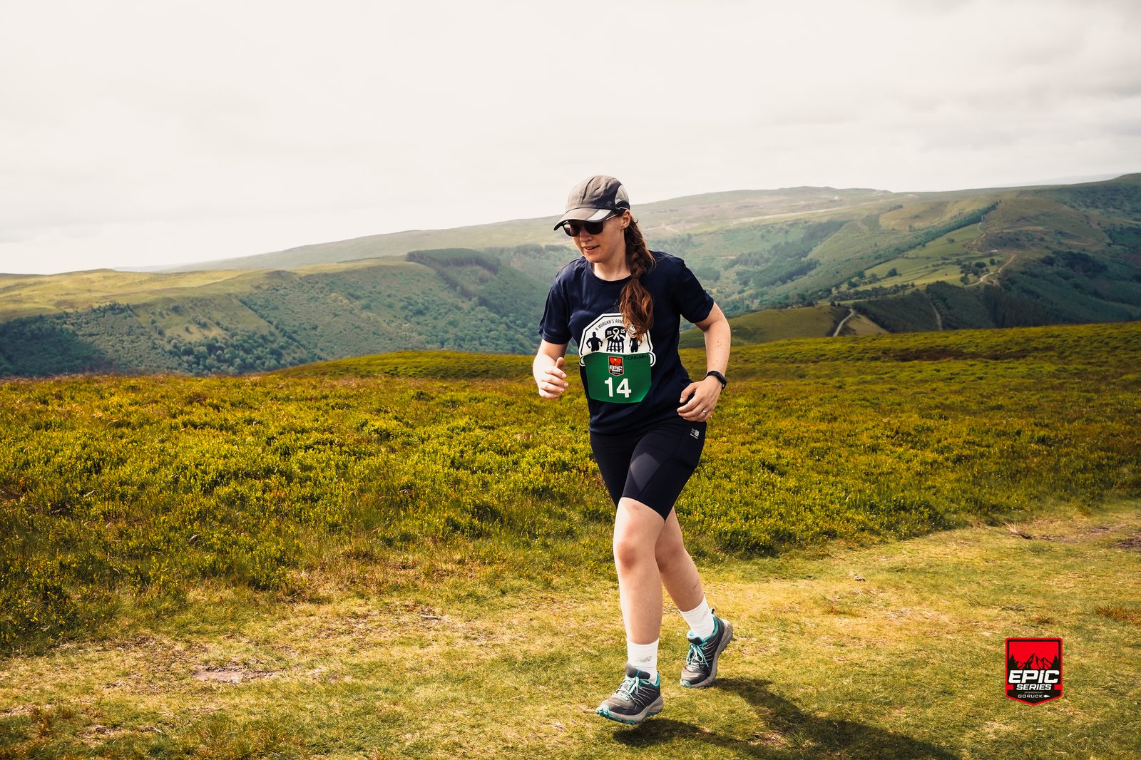 A person wearing sunglasses, a cap, and race number 14 runs on a grassy trail with green hills and valleys in the background under a cloudy sky. A small "EPIC" event logo appears in the bottom right corner.