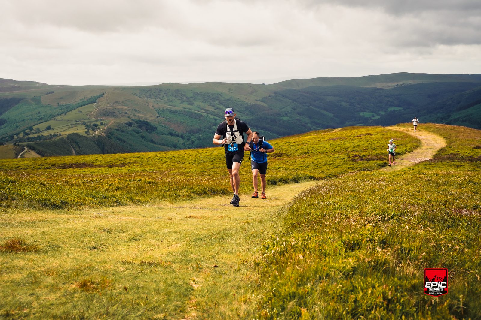 Several trail runners race along a scenic path on a grassy hillside, with rolling green mountains in the background under a cloudy sky. A red "Epic" race sign appears in the bottom right corner.