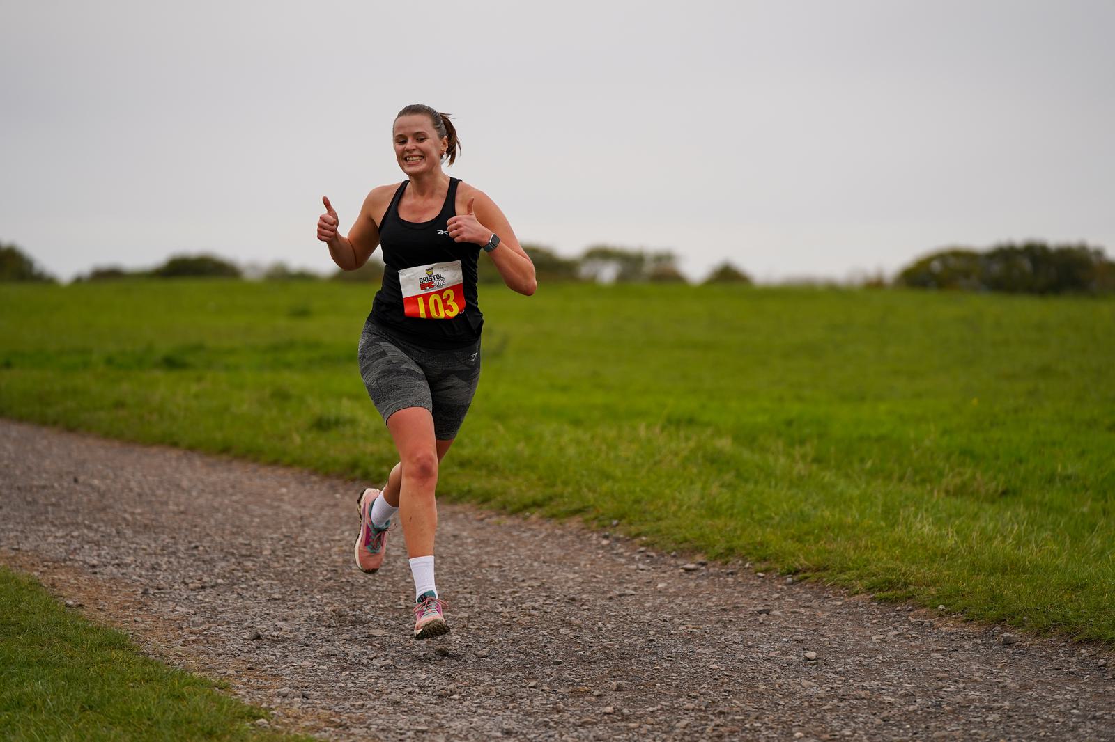 A person in athletic gear running on a gravel path through a grassy field, giving a thumbs-up. They have a numbered race bib on their shorts, and the sky is overcast.