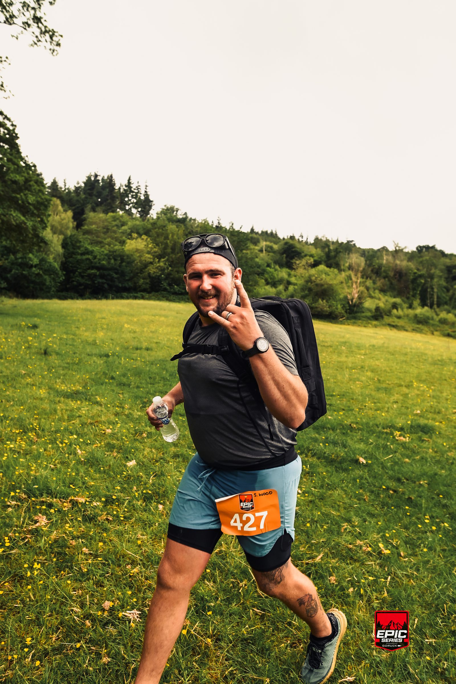 A man wearing athletic gear, sunglasses, and a backpack smiles and flashes a peace sign while walking through a grassy field with trees in the background. He has a race bib numbered 427 and holds a water bottle.