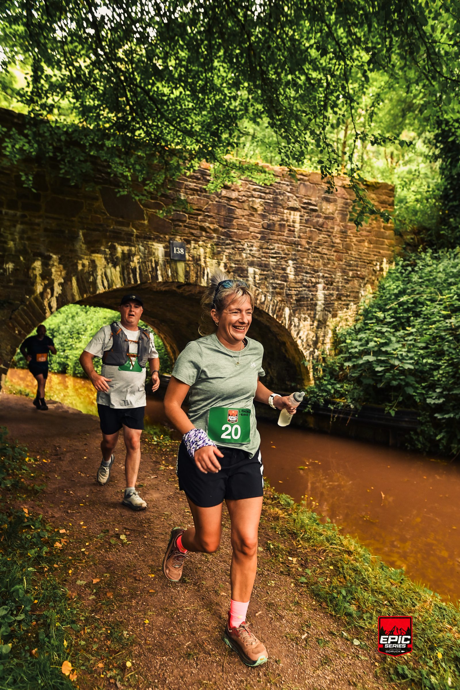Two runners, a woman smiling in front and a man behind, jog along a canal path under a stone bridge surrounded by lush greenery during a trail race event.