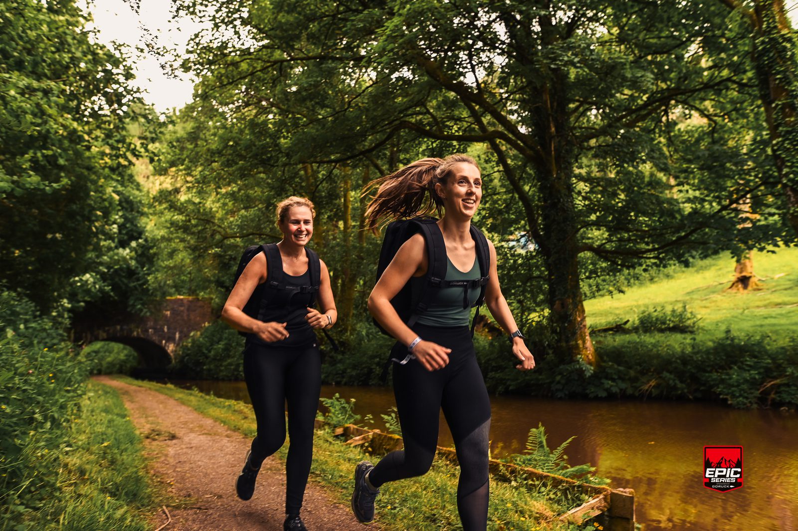 Two women in athletic gear smile while trail running on a dirt path beside a canal, surrounded by lush green trees, with a stone bridge and an "Epic" event logo in the background.