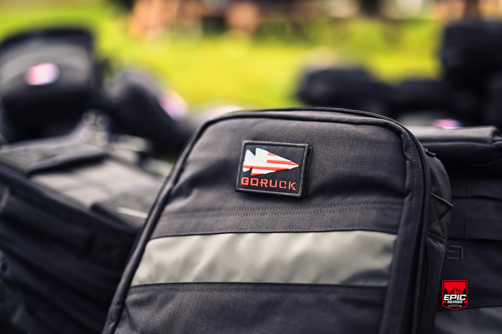 Close-up of a black backpack with a GORUCK patch on the front. Other backpacks are visible in the blurred background, and the scene appears to be outdoors with greenery.