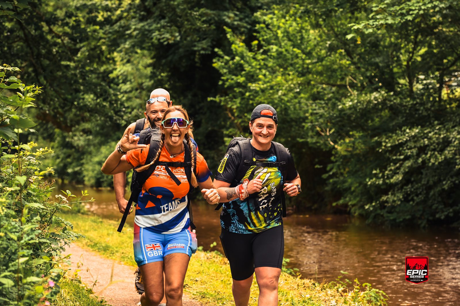 Three athletes in outdoor gear run beside a river through a lush, green forest trail. The front runner smiles and gestures at the camera, while the others follow close behind. A red "EPIC" event logo is visible in the bottom right corner.