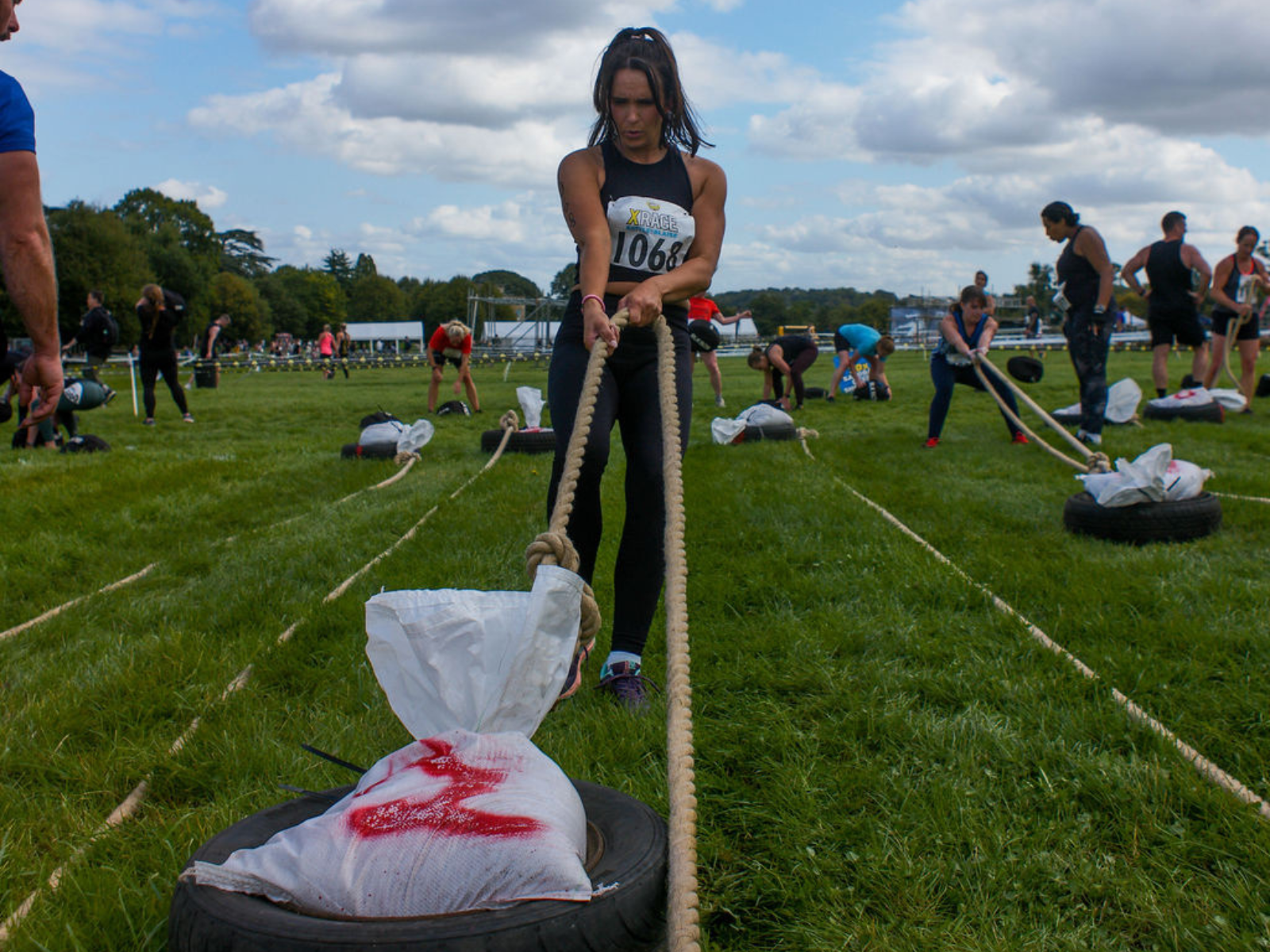 A woman competes in an outdoor strength challenge, pulling a heavy weight attached to a rope. Other participants are seen in the background, also engaging in similar activities on a grassy field under a cloudy sky.