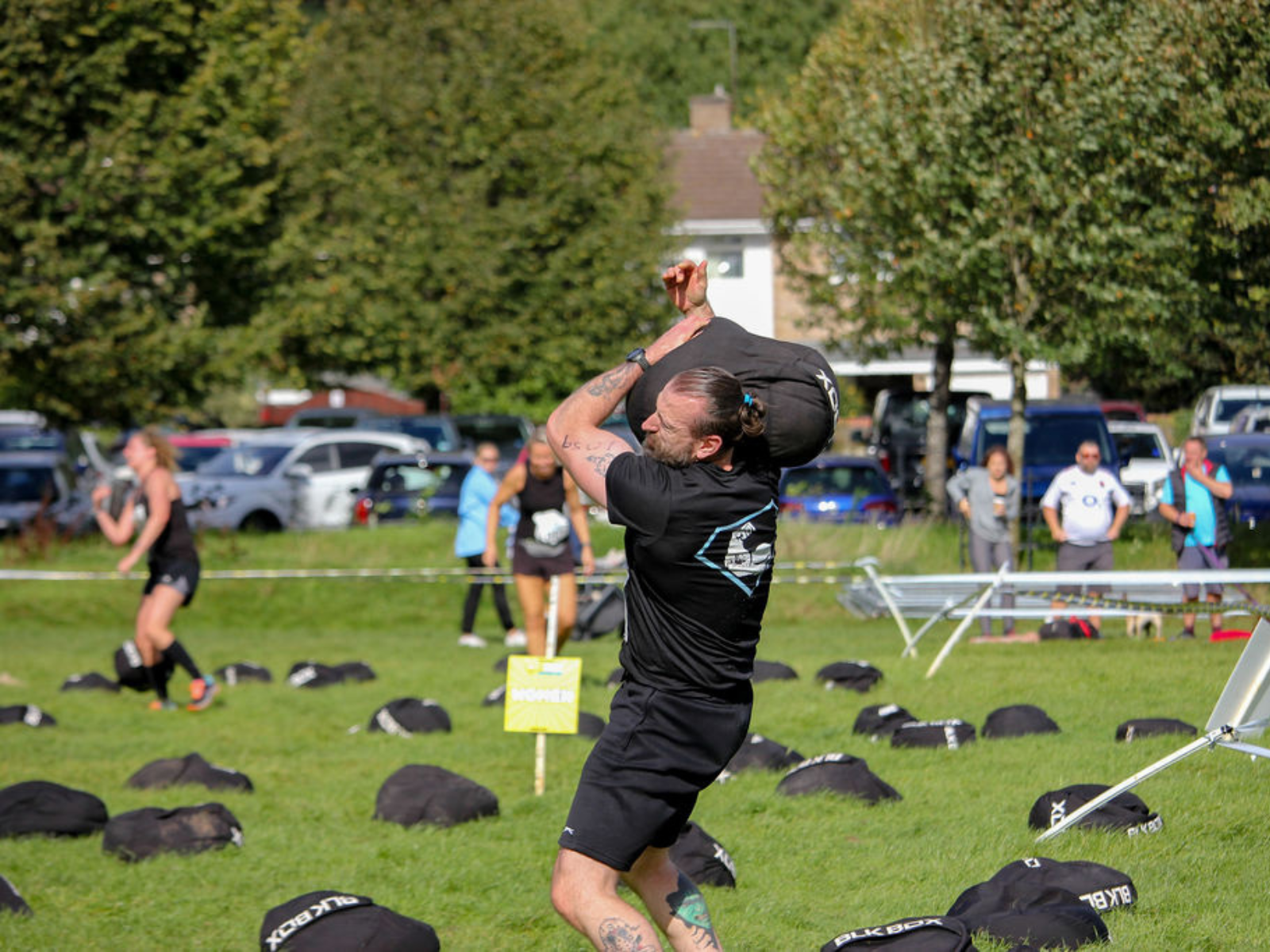 A man in a black athletic outfit lifts a sandbag above his shoulders during an outdoor fitness competition. Other participants and spectators are in the background, with cars parked near a tree-lined area. The ground is dotted with more sandbags.