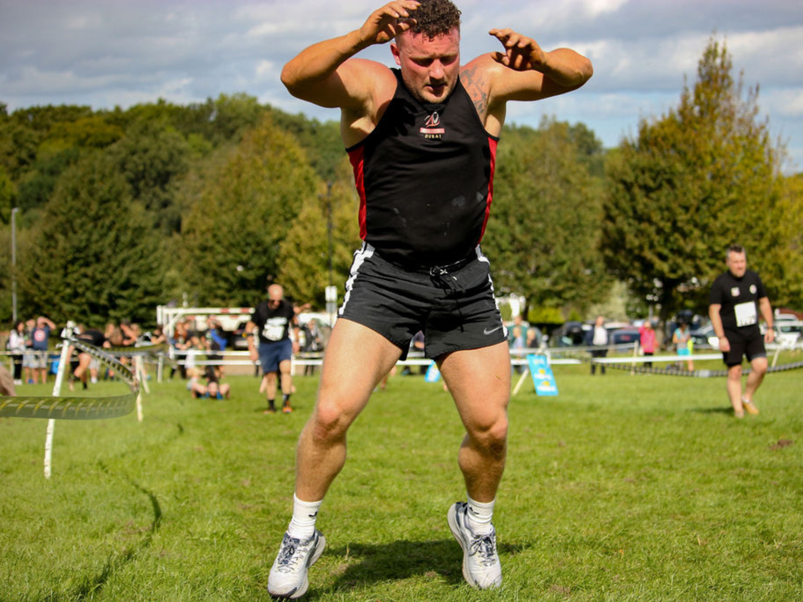 A man with short hair in a black tank top and shorts is mid-air during an outdoor athletic event, possibly a jump. He has his arms raised and legs bent. The background shows other participants, trees, and a partly cloudy sky.
