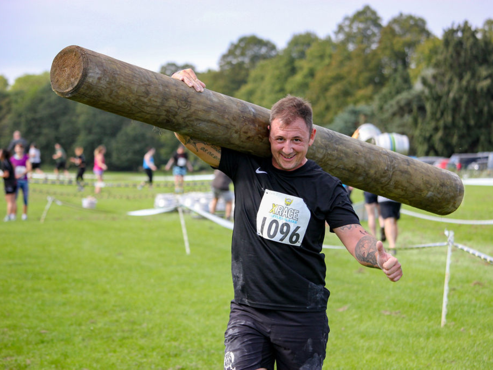 A person wearing a black shirt and black shorts is smiling and giving a thumbs up while carrying a large log over their shoulder during an outdoor event. The background shows grass, other participants, trees, and event barriers.