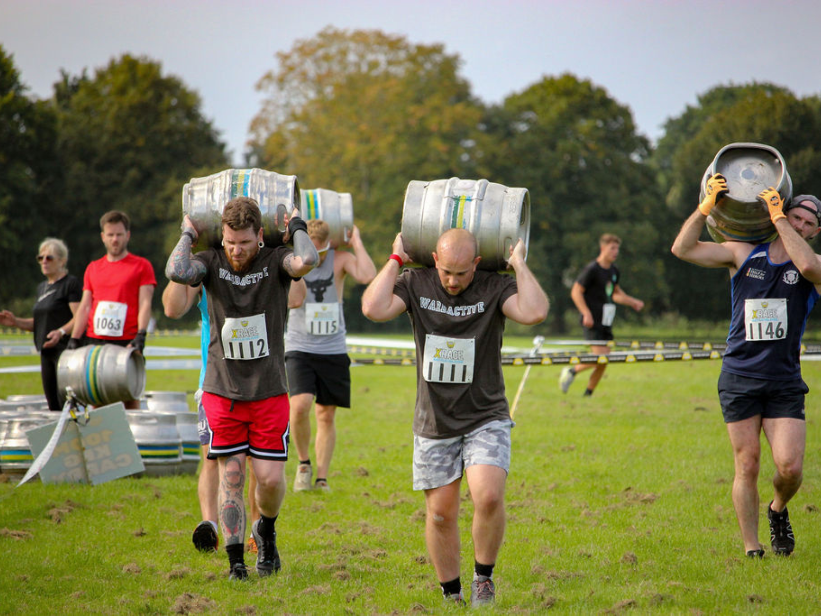 Participants in an outdoor competition carry metal kegs on their shoulders across a grassy field. They wear casual athletic attire and numbered race bibs. Other competitors and trees are visible in the background.