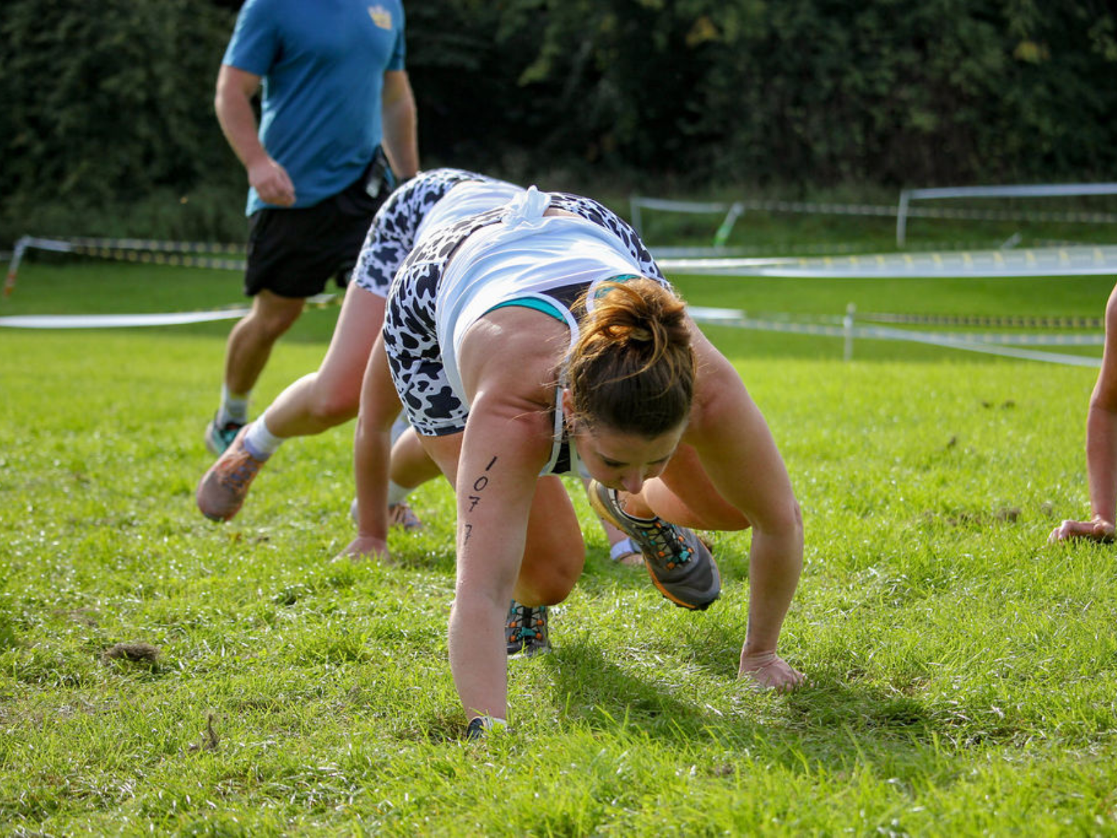 A woman participating in an outdoor obstacle course, crawling on the grass wearing athletic gear. Her arm has a visible number written on it. Other participants and parts of the course are visible in the background.