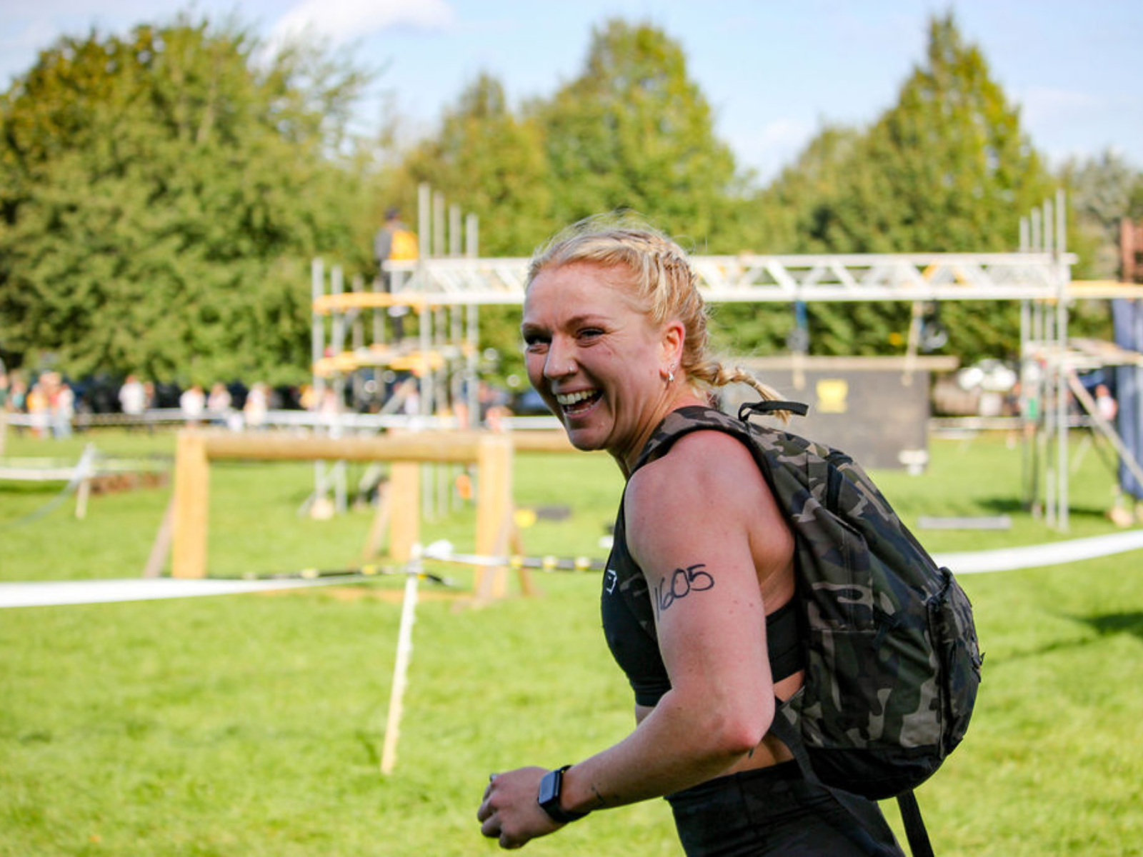A woman, wearing athletic gear and carrying a camouflage backpack, smiles while participating in an outdoor event. She has "205" written on her upper arm and appears to be in the middle of an obstacle course on a grassy field with trees and structures in the background.