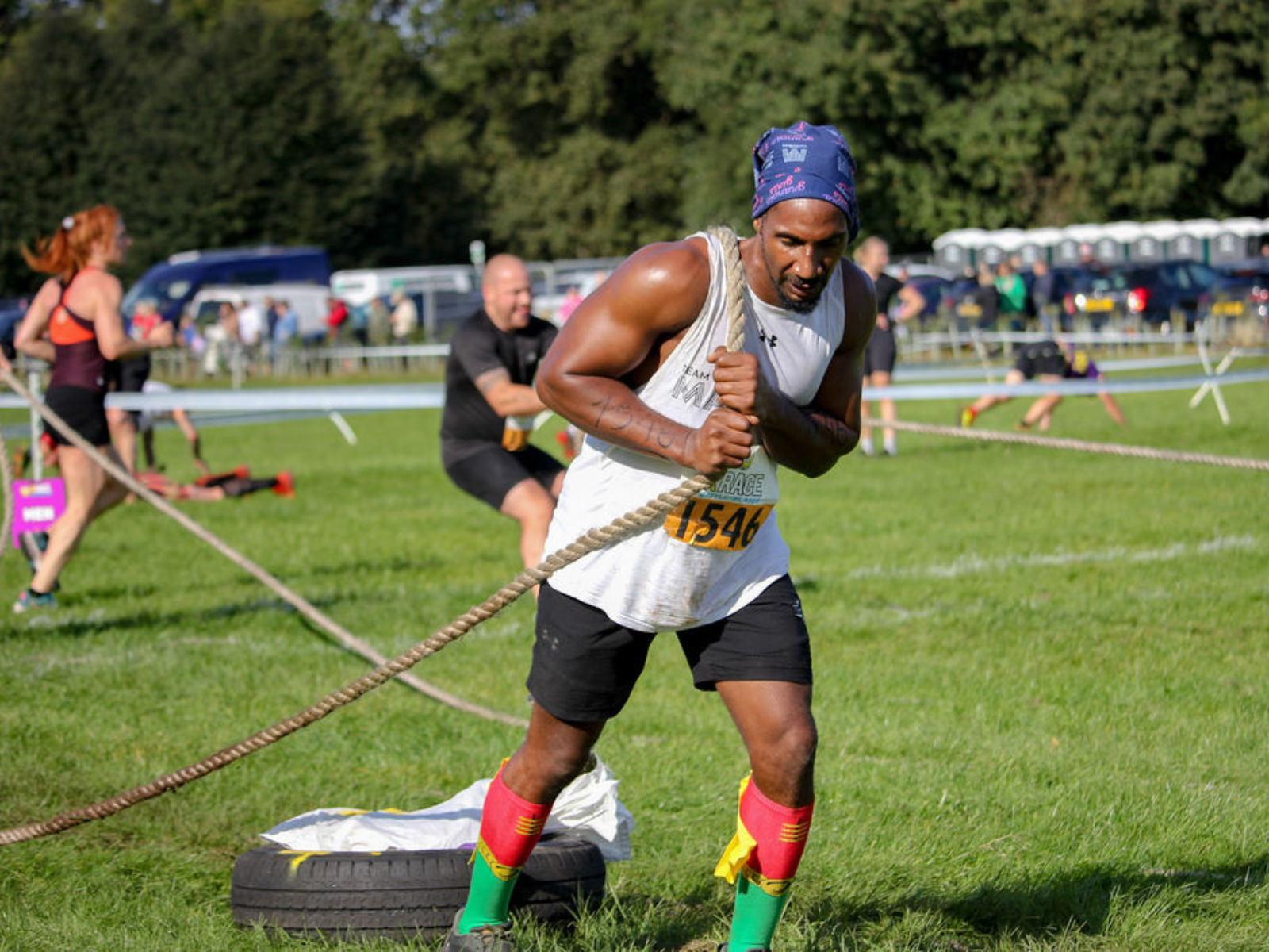 A man wearing a white tank top, black shorts, red socks, and a bandana pulls a heavy rope attached to a tire during an outdoor fitness event. Other participants are visible in the background engaged in similar activities. It's a sunny day on a grassy field.