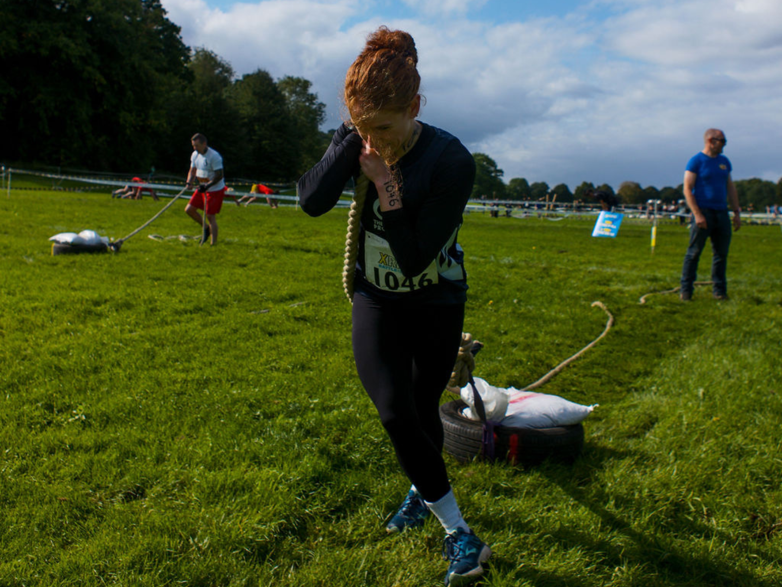 A person with curly hair is pulling a tire with sandbags tied to it across a grassy field during an outdoor competition. The individual looks focused and determined. Other competitors and spectators are visible in the background.