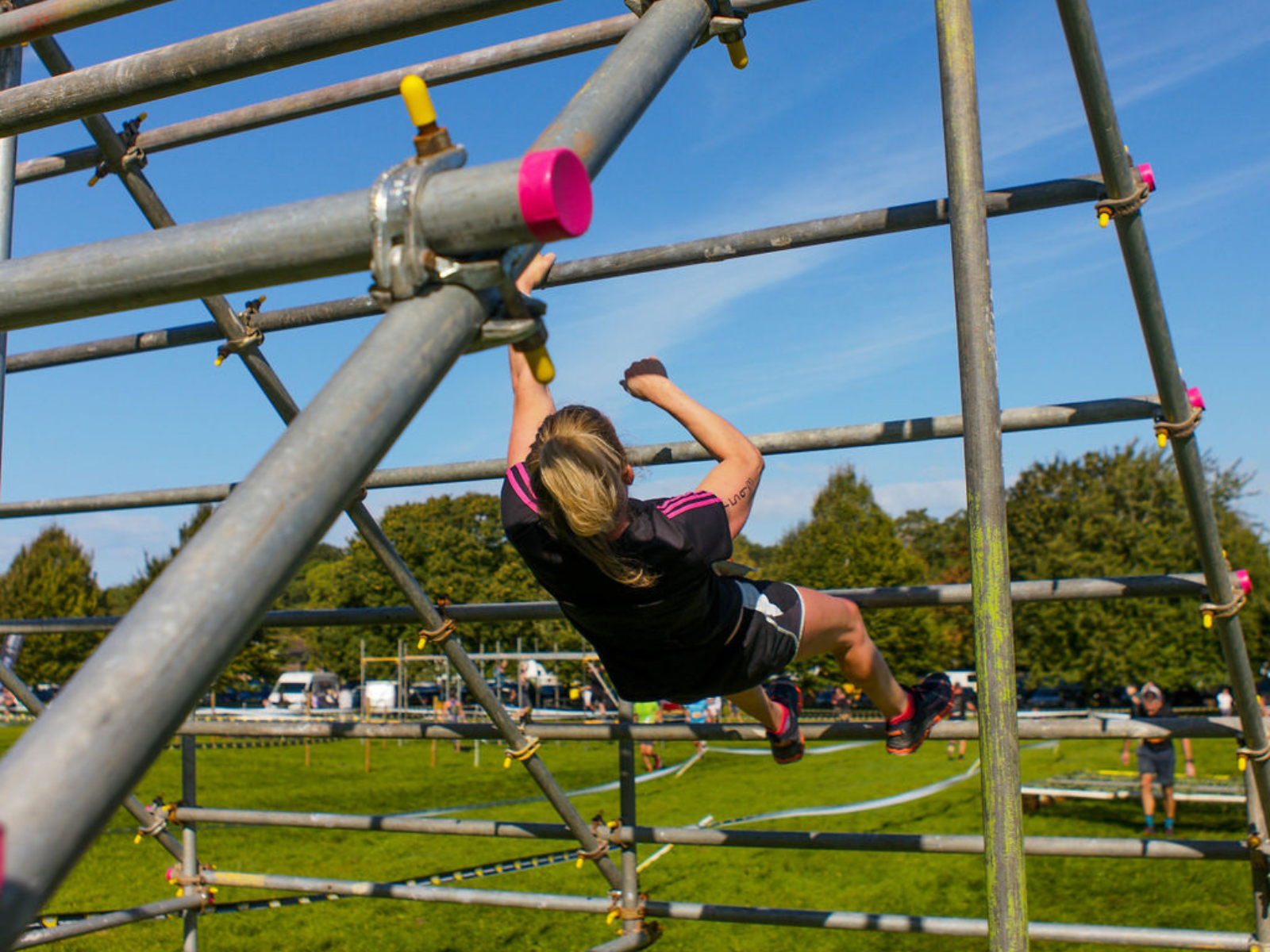 A person with blonde hair wearing athletic gear is climbing on a metal frame structure outdoors. The sky is blue, and green grass and trees can be seen in the background. Other participants and additional obstacles are visible in the distance.