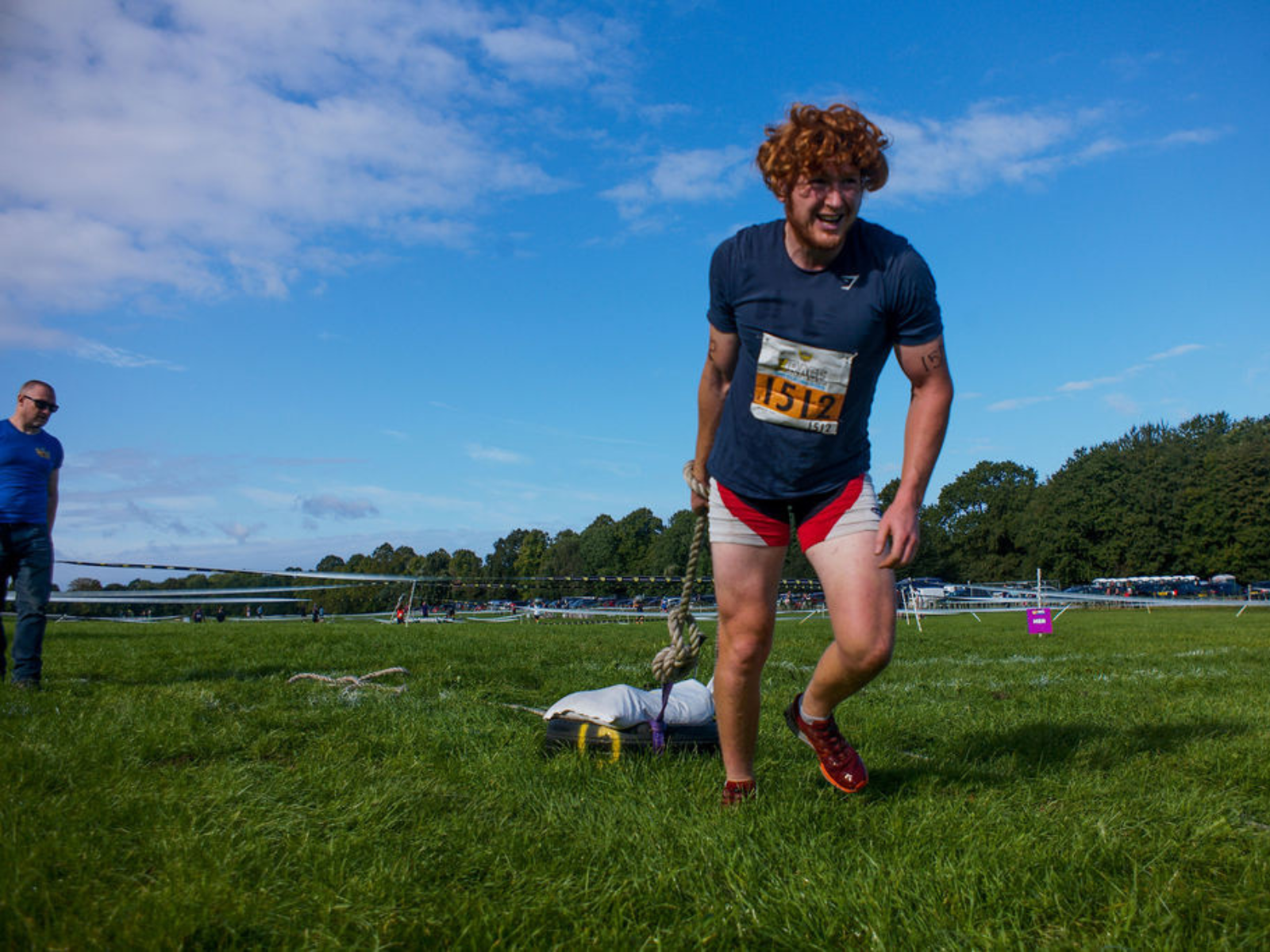 A person with curly hair, wearing a race bib, pulls a weighted object across a field during an outdoor event. They are wearing a dark T-shirt, red shorts, and red shoes. The sky is partly cloudy, and several people and trees are visible in the background.